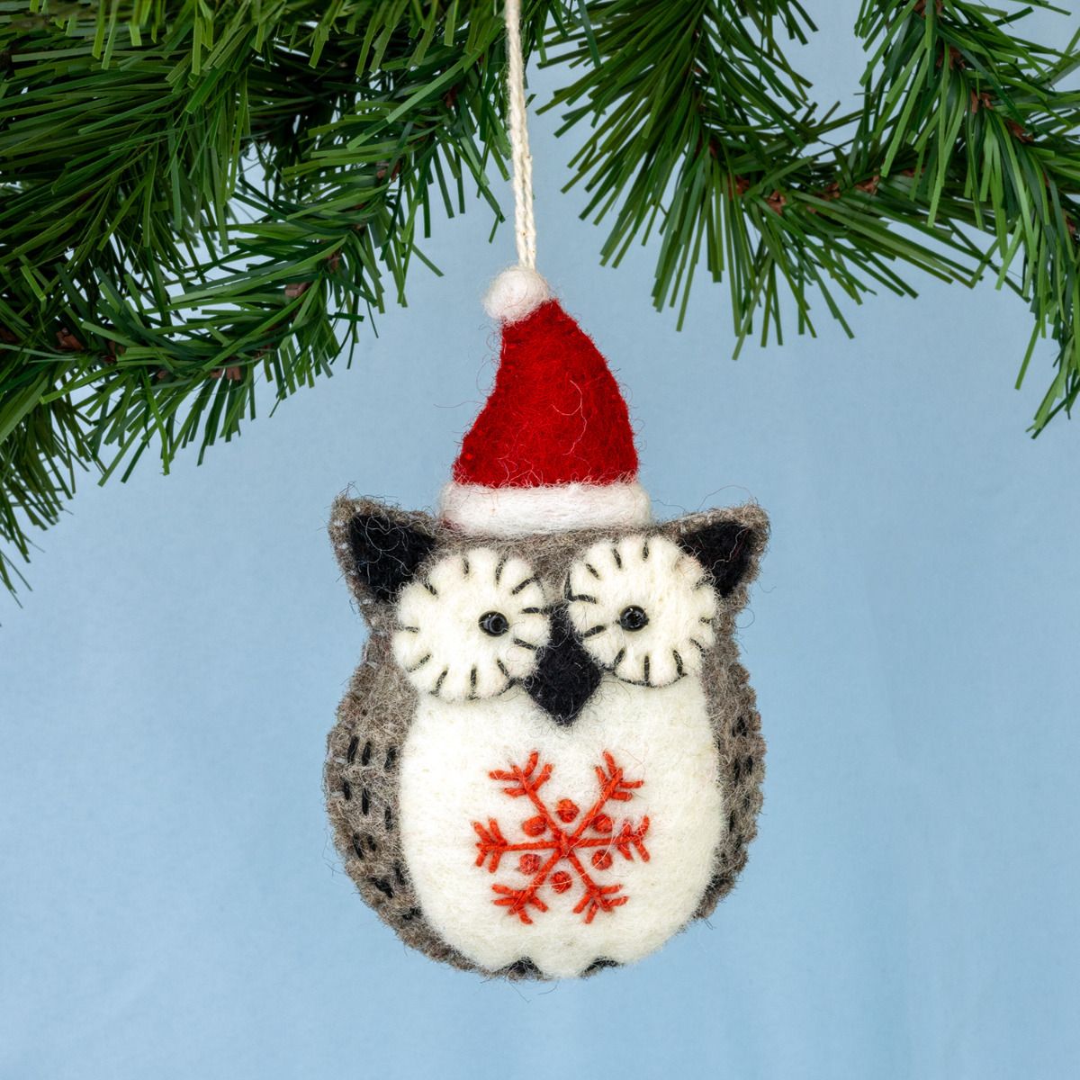 Felt owl ornament with a red snowflake design and Santa hat hanging on a Christmas tree.