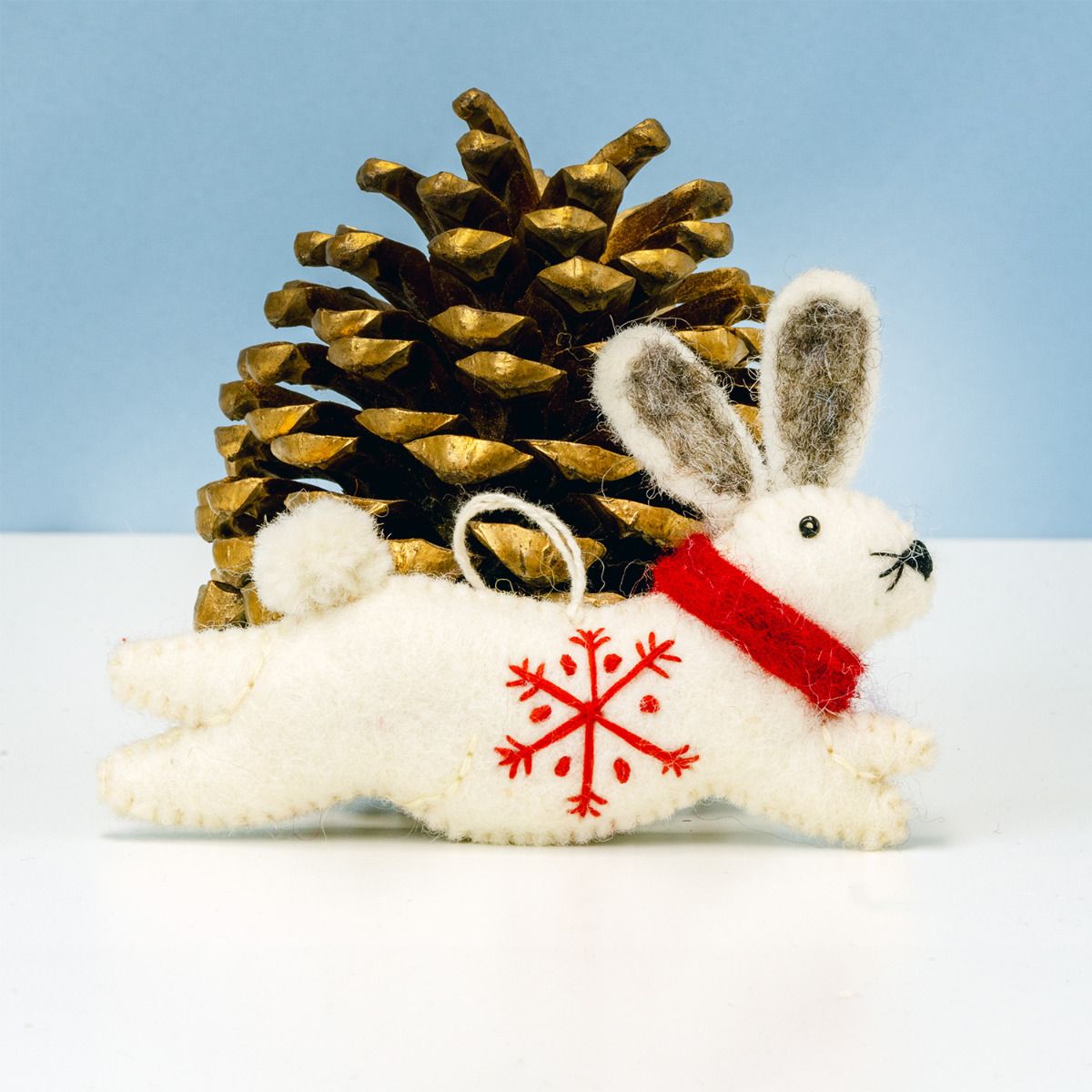 White rabbit ornament with red snowflake design and red scarf next to a gold pine cone on a light blue background.
