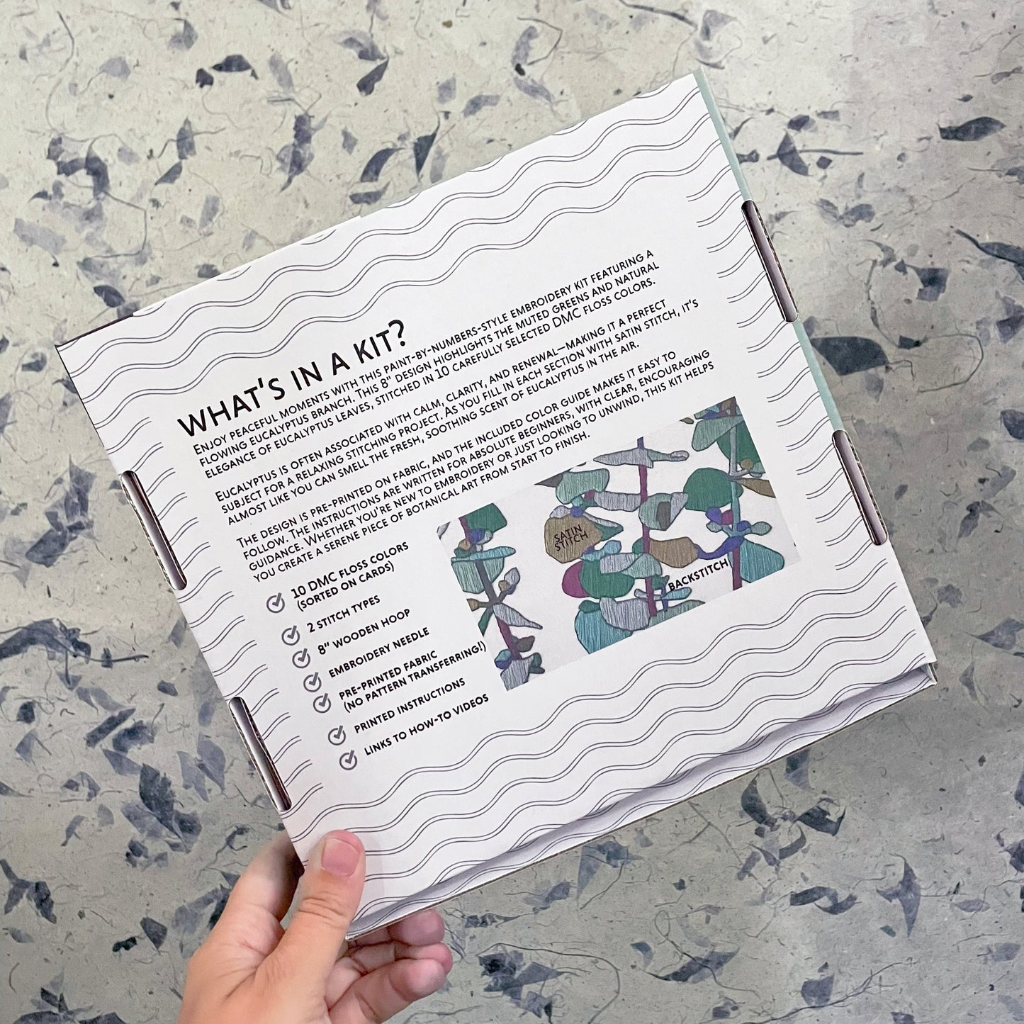 Hand holding a craft kit packaging with text and illustrations on a textured surface