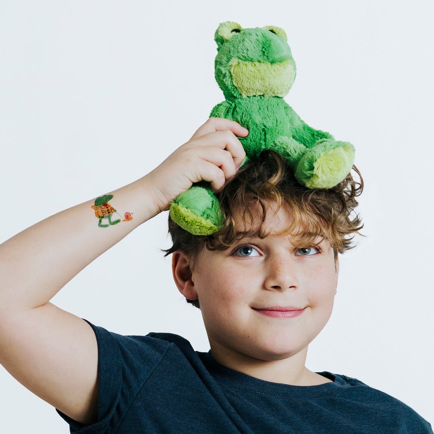 Child with a green plush frog toy on their head against a white background. The child wears a temporary tattoos of a frog in a raincoat, walking its pet snail on a leash.