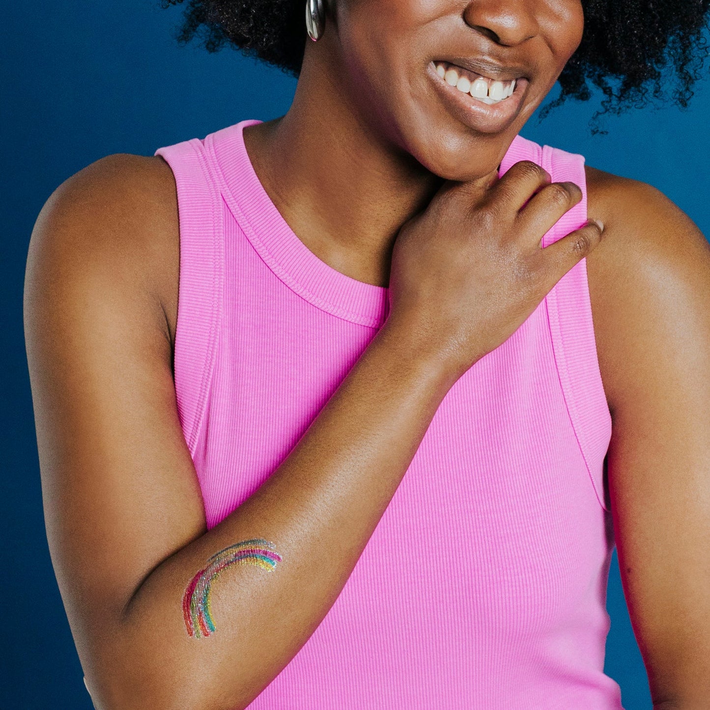 Woman wearing a pink tank top with a colorful rainbow tattoo on her arm against a blue background