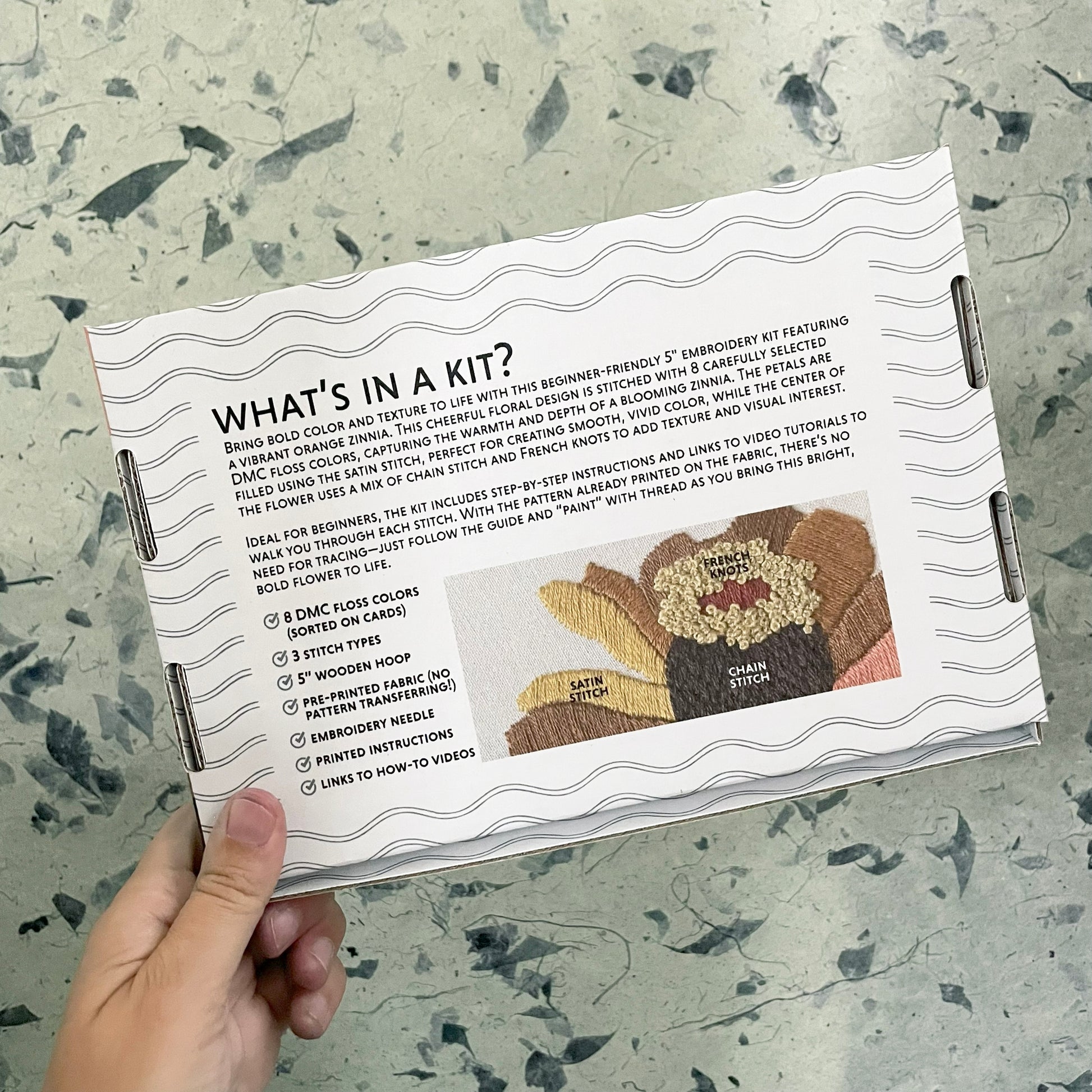 Hand holding a craft kit with an embroidered turkey design and text on a textured surface.