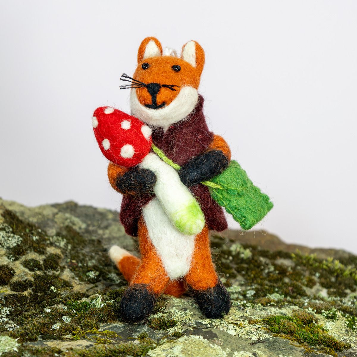 Felted fox ornament holding a red mushroom on a mossy surface