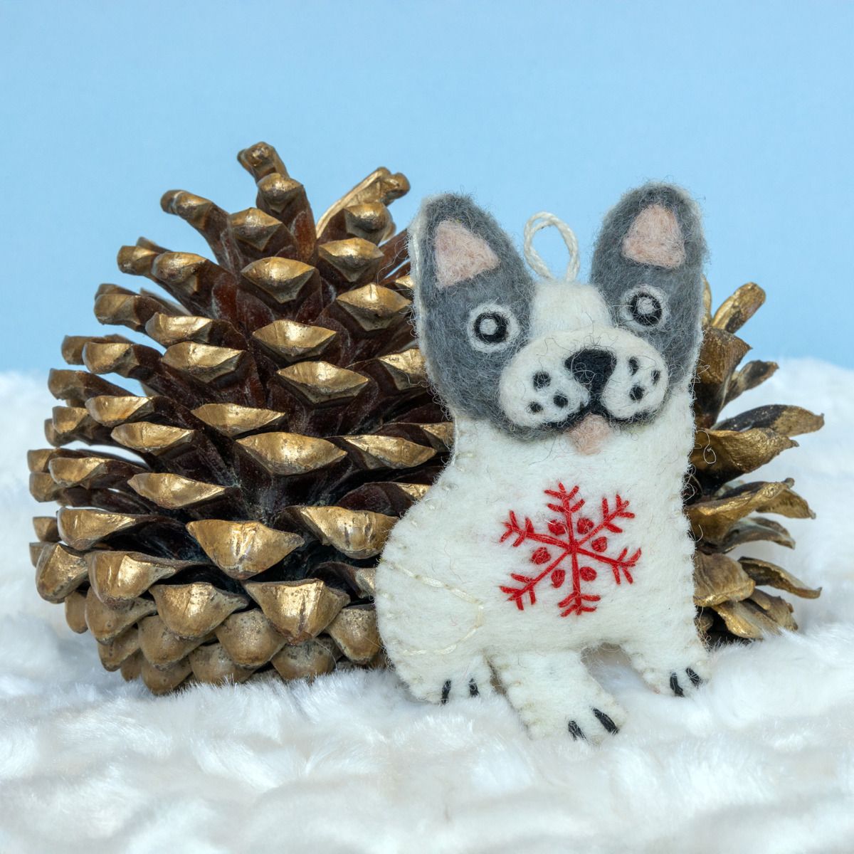 Small plush dog ornament with a red snowflake design next to a large pine cone on a snowy background