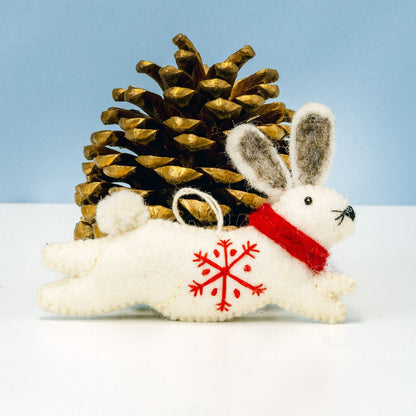 White rabbit ornament with red snowflake design and red scarf next to a gold pine cone on a light blue background.