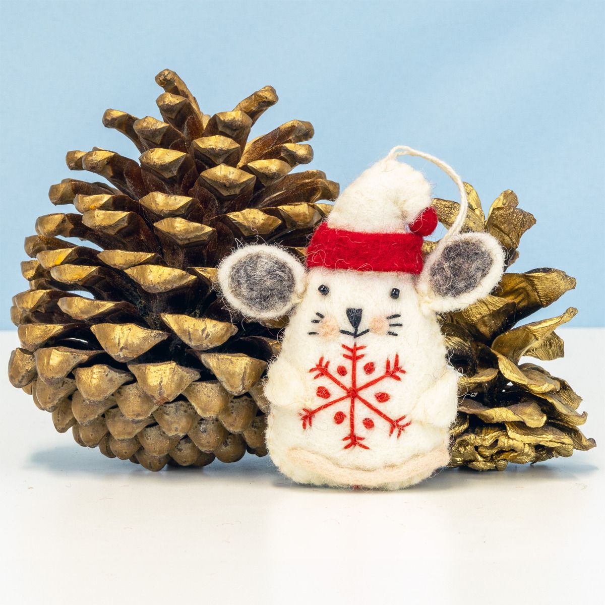 Decorative mouse with red snowflake pattern and red scarf next to a pine cone on a light blue background