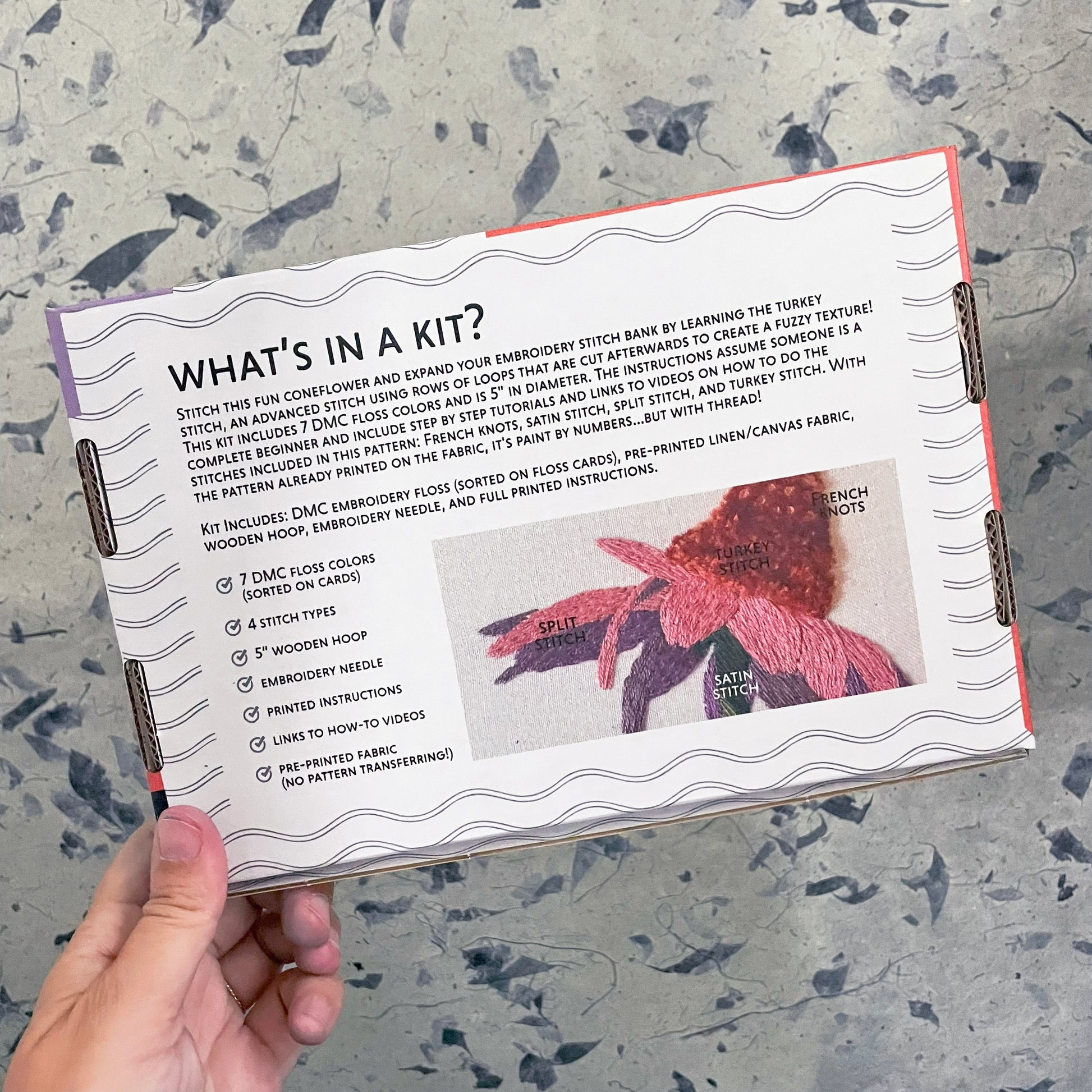 Embroidery kit information sheet held by a hand on a textured surface