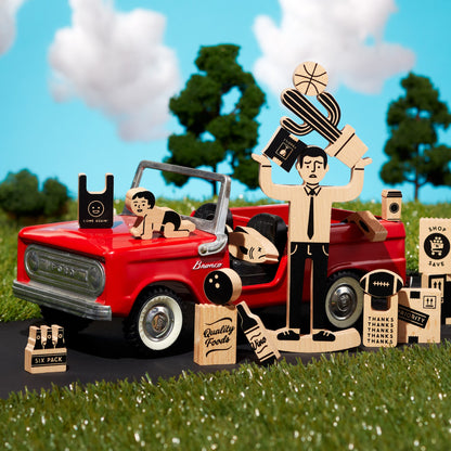 Wooden toy set with a red truck, people, and various items on grass with a blue sky background