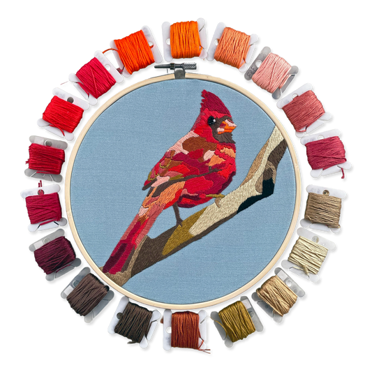 Embroidery hoop with a red cardinal bird design surrounded by colorful threads on a white background