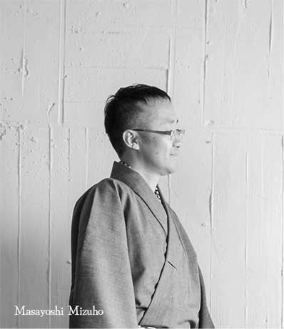 Black and white portrait of a person wearing glasses and a suit against a light wooden panel background.