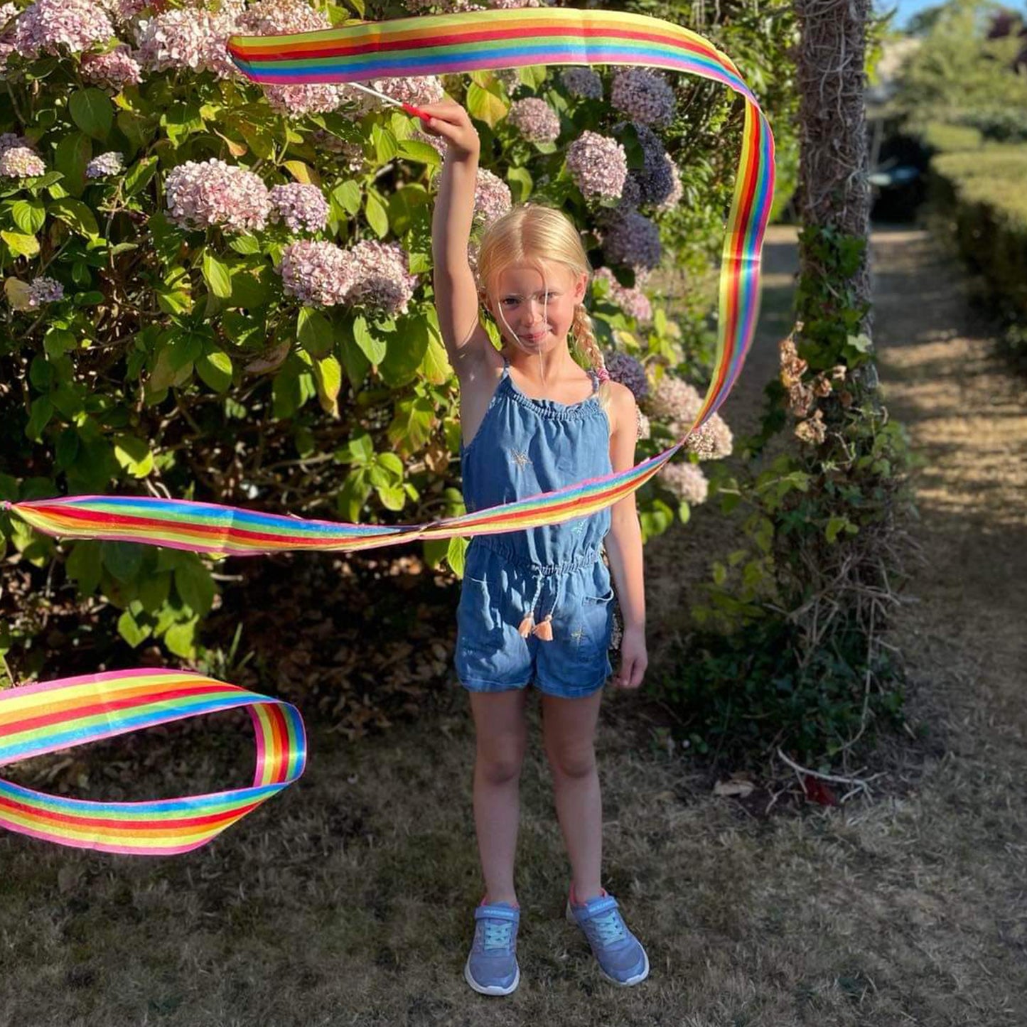 Young girl holding a long rainbow ribbon in an outdoor setting with flowers and trees.