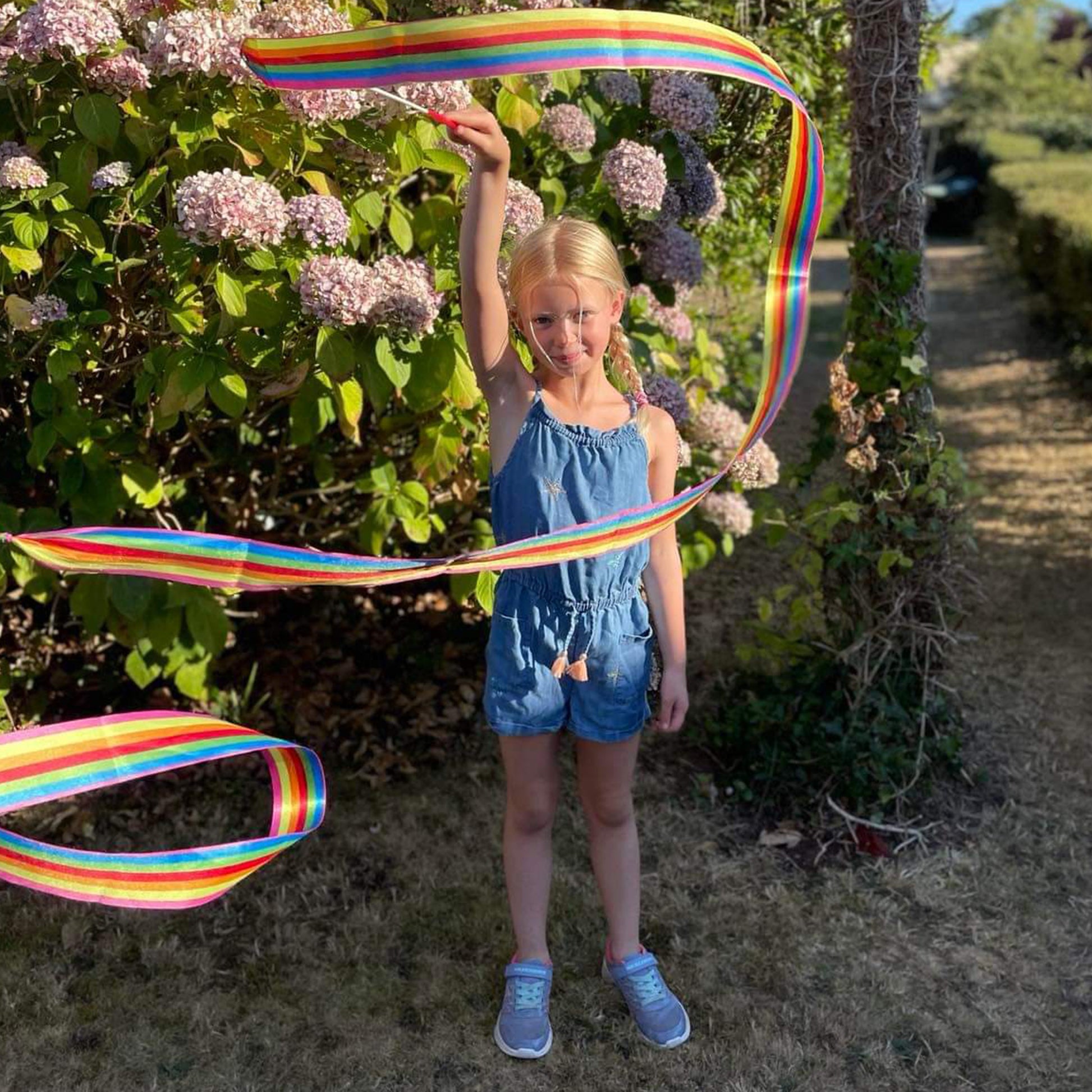 Young girl holding a long rainbow ribbon in an outdoor setting with flowers and trees.