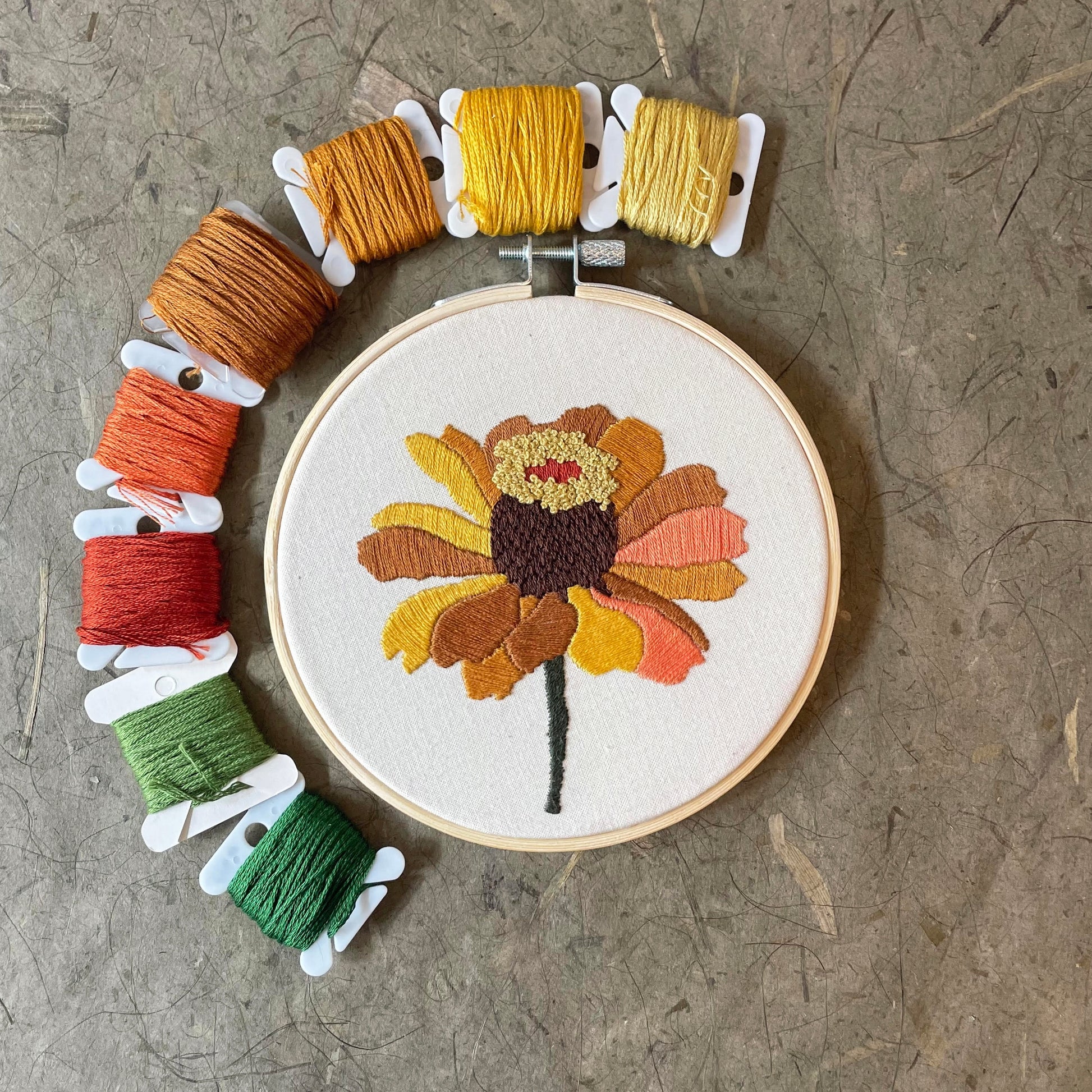 Embroidery hoop with a colorful floral design surrounded by spools of thread on a textured surface.