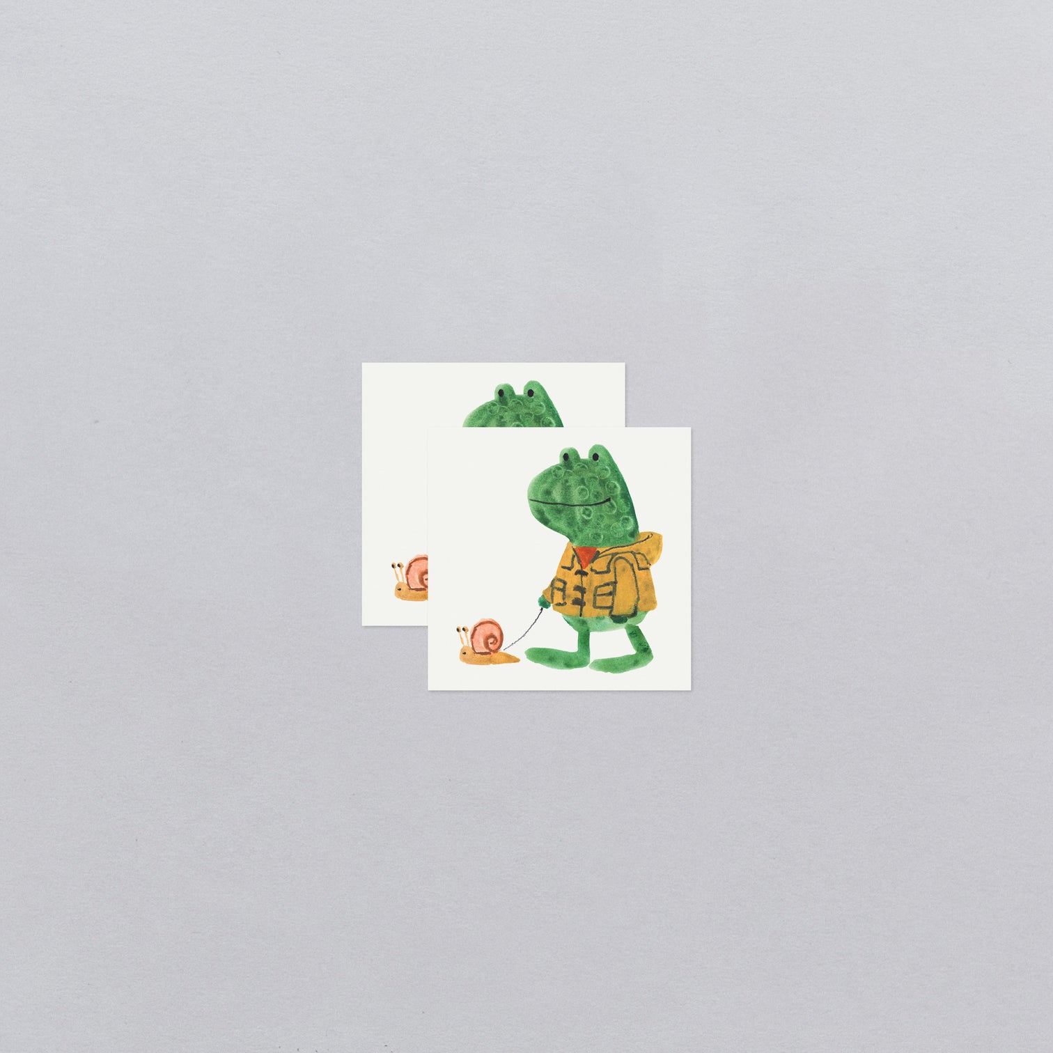 Two temporary tattoos of a green frog character with a yellow raincoat on a gray background